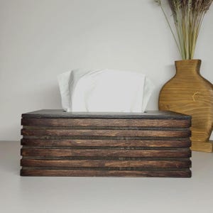 May include: A rectangular tissue box cover constructed from dark brown stained wood with horizontal slats. The box features a smooth top surface with a central rectangular opening, through which a stack of white facial tissues is dispensed.