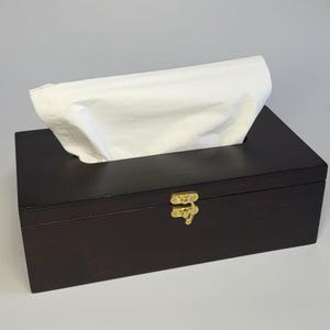 May include: A rectangular dark brown wooden tissue box holder with a hinged lid and a gold-colored metal latch on the front. A single white paper tissue is pulled through the rectangular opening on the top of the box.