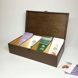 Könnte beinhalten: Eine rechteckige Teebox aus dunkelbraunem Holz mit Klappdeckel und Metallverschluss. Das Innere ist in vier Fächer unterteilt, die jeweils Reihen von Teebeuteln in weißer, grüner und violetter Verpackung mit verschiedenen Aufdrucken enthalten.