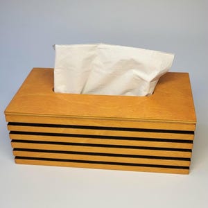 May include: A rectangular wooden tissue box cover featuring a light brown finish with horizontal slats separated by thin black gaps. A single white paper tissue is pulled through the rectangular opening on the top surface of the wooden container.