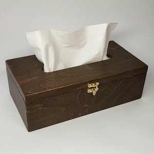 May include: A rectangular dark brown wooden tissue box holder with a visible wood grain pattern. The box features a gold-coloured metal latch on the front and a rectangular opening on the top, from which a single white paper tissue is protruding.
