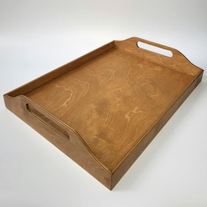 May include: A rectangular wooden serving tray with a smooth, medium-brown finish and visible wood grain patterns. The tray features raised sides and two integrated cutout handles on the shorter ends for easy carrying and handling.