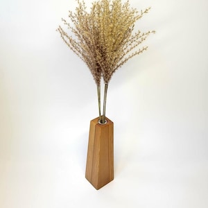 Könnte beinhalten: Eine hohe, konisch zulaufende Holzvase mit hellbrauner Maserung und einem kreisförmigen Metallrand an der oberen Öffnung. Die Vase enthält ein Bündel getrockneter, federartiger beiger Gräser, die sich nach oben hin auffächern und ein Arrangement bilden.
