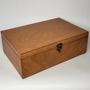 May include: A rectangular wooden box with a medium brown finish and visible wood grain patterns. The box features a hinged lid and a small, antique-style bronze-colored metal latch centered on the front edge to secure the closure.