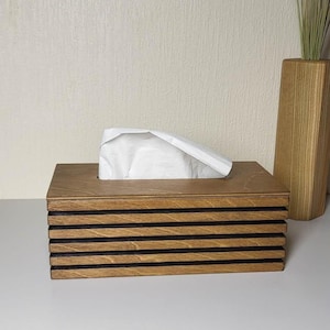 Pode incluir: Uma caixa de lenços retangular de madeira com um design em camadas de ripas de madeira castanho-claro alternadas com finas riscas horizontais pretas. Um lenço de papel branco sobressai da abertura na parte superior.