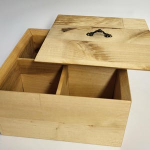 May include: A rectangular light-colored wooden storage box with a natural grain pattern. The box features an open interior divided into two sections by a central partition. A matching wooden lid with a small dark metal handle rests partially open.