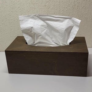 May include: A rectangular tissue box cover made of dark brown stained wood with a visible natural grain pattern. A single white paper tissue is pulled through the oval-shaped opening at the top, extending upwards from the wooden container.