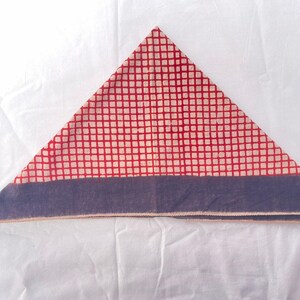 May include: A triangular folded fabric piece featuring a red and off-white grid pattern across the main body. The base of the triangle is finished with a solid dark blue horizontal border that runs along the entire bottom edge of the textile.