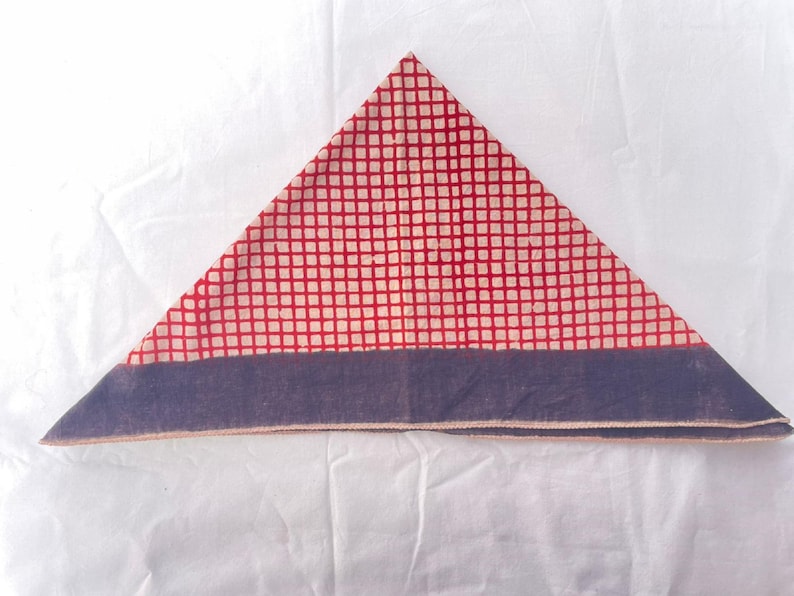 May include: A triangular folded fabric piece featuring a red and off-white grid pattern across the main body. The base of the triangle is finished with a solid dark blue horizontal border that runs along the entire bottom edge of the textile.