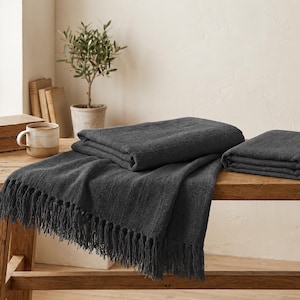 May include: Two folded charcoal grey blankets with a textured weave and long fringe edges. The blankets are displayed on a wooden surface, showcasing a deep, dark grey colour and a consistent, woven fabric pattern throughout the textile pieces.