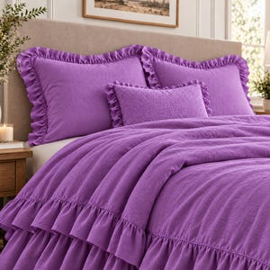 Puede incluir: Un juego de cama de color morado vibrante que incluye dos almohadas rectangulares grandes con bordes de volantes, un cojín rectangular pequeño con textura y un edredón a juego con varias capas de volantes en el borde inferior.