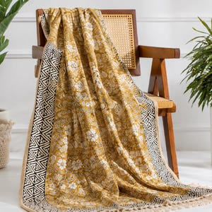 Puede incluir: Una manta de color amarillo mostaza con un estampado floral blanco y bordes con diseños geométricos en blanco y negro. La pieza de tela rectangular tiene flecos en los extremos y está colocada sobre una silla de madera con respaldo de rejilla.