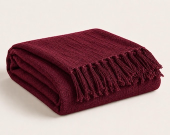 May include: A folded deep burgundy throw blanket featuring a textured knit pattern and a row of matching fringe along the edge. The fabric has a consistent dark red colour and a woven appearance, neatly stacked in a rectangular shape.