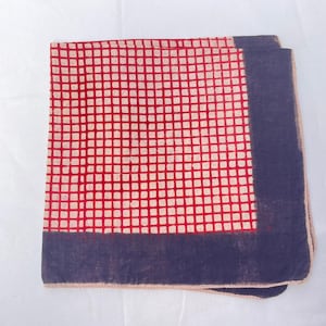 May include: A square fabric handkerchief featuring a central grid pattern of red lines on a cream background, bordered by a solid dark blue band along two edges. The textile has a visible weave and a finished hem along the outer perimeter.