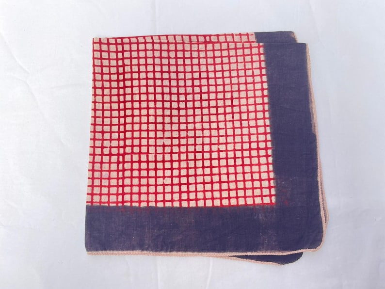 May include: A square fabric handkerchief featuring a central grid pattern of red lines on a cream background, bordered by a solid dark blue band along two edges. The textile has a visible weave and a finished hem along the outer perimeter.