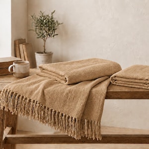 May include: Two folded tan blankets with a textured weave and long fringe trim. The blankets are displayed on a rustic wooden table alongside a ceramic mug and a stack of books, creating a neutral-toned arrangement with a visible fabric weave.