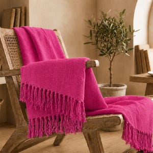 Könnte beinhalten: Eine leuchtend pinkfarbene Wohndecke mit strukturierter, gerippter Oberfläche und langen, geknoteten Fransen an den Rändern. Die Decke liegt über einem Holzstuhl mit einem Rückenlehnen-Einsatz aus Wiener Geflecht und zeigt ihren weichen Stoff.
