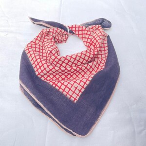 May include: A triangular bandana featuring a central cream-colored section with a red grid pattern. The edges are bordered by a solid dark blue fabric, and the entire piece is tied at the top, creating a folded, layered appearance with visible stitching.