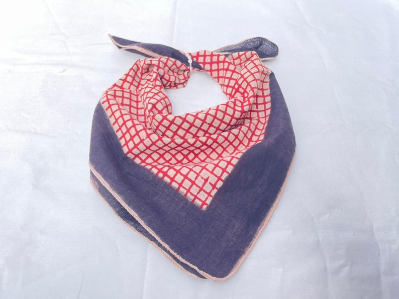 May include: A triangular bandana featuring a central cream-colored section with a red grid pattern, bordered by a solid dark blue fabric. The edges of the fabric are finished with a light-colored hem, and the top corners are tied in a knot.
