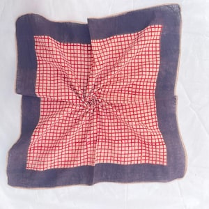 May include: A square handkerchief featuring a central grid pattern of red lines on a cream background, framed by a wide, solid dark purple border. The fabric is folded toward the center, creating a subtle spiral effect in the textile weave.