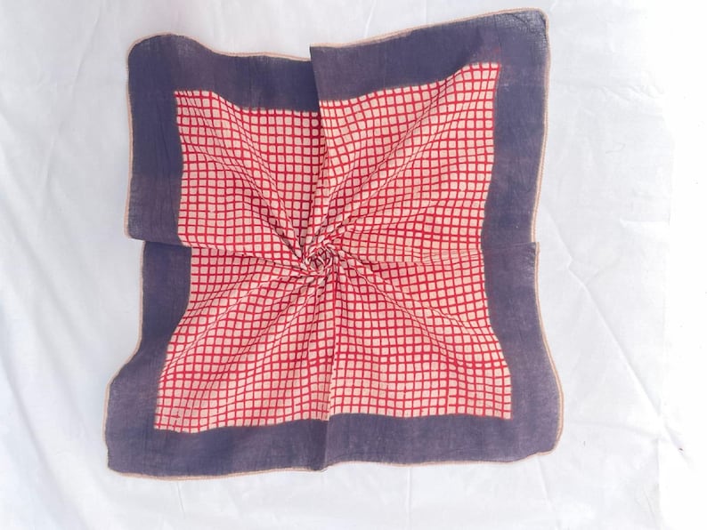 May include: A square handkerchief featuring a central grid pattern of red lines on a cream background, framed by a wide, solid dark purple border. The fabric is folded toward the center, creating a subtle spiral effect in the textile weave.