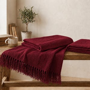 May include: A deep burgundy throw blanket with a textured weave and long fringe along the edges. The blanket is folded into two separate pieces, one draped over the other, showcasing the rich, dark red color and the soft, woven fabric construction.