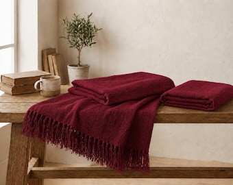 May include: A deep burgundy throw blanket with a textured weave and long fringe along the edges. The blanket is folded into two separate pieces, one draped over the other, showcasing the rich, dark red colour and the soft, woven fabric construction.