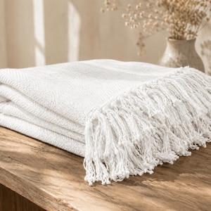 May include: A folded white throw blanket with a textured woven pattern and long, knotted fringe trim along the edge. The fabric has a visible weave structure, and the fringe consists of multiple thin, hanging strands of white yarn.