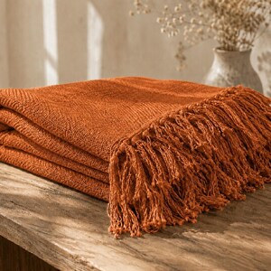 May include: A folded burnt orange throw blanket featuring a textured weave pattern and long, decorative fringe along the edge. The fabric has a consistent, deep rust-colored hue and a visible, slightly coarse textile construction throughout the piece.