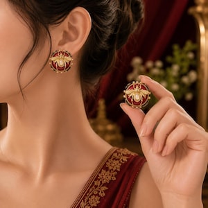 May include: A pair of round stud earrings featuring a gold-toned bee motif centred on a deep red circular base. The bee has an oval white stone at its centre and is surrounded by a beaded gold border. One earring is worn on the earlobe.