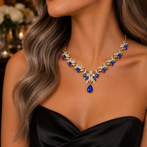 May include: A gold-toned necklace featuring a series of marquise-cut blue stones and clear crystals arranged in a leaf-like pattern. The centrepiece includes a prominent teardrop-shaped blue stone hanging from a floral-inspired metal setting.