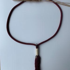 Könnte beinhalten: Eine kreisförmige, dunkel kastanienbraune Kordelkette mit einer langen, passenden Quaste, die an einem cremefarbenen, umwickelten Akzent hängt. Ein kleiner metallischer Blattanhänger ist an der Oberseite der Quaste befestigt. Drei getrocknete beige Grashalme liegen darüber.