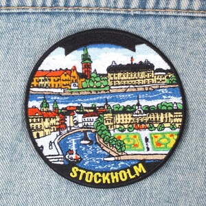 May include: A circular embroidered patch featuring a colorful cityscape illustration of Stockholm, Sweden. The design includes buildings, a river, and green spaces, with the word STOCKHOLM stitched in yellow capital letters at the bottom edge.