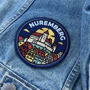 May include: A circular embroidered patch featuring a stylised illustration of the Nuremberg castle and city skyline against a yellow sun, with the text NUREMBERG in white letters, attached to a light blue denim jacket with a metal button.
