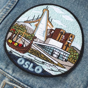 May include: A circular embroidered patch featuring a stylised depiction of Oslo, Norway, including a brown Viking ship, a tall white tower, and modern buildings. The word OSLO is stitched in light blue letters at the bottom against a dark blue background.