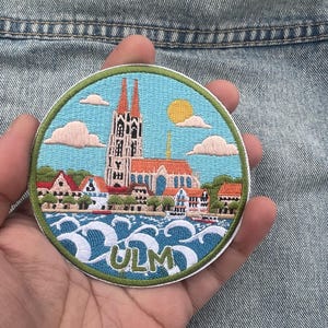 May include: A circular embroidered patch featuring a detailed scene of the Ulm Minster cathedral with its twin spires, surrounded by houses, trees, and stylized blue waves. The word ULM is stitched in green at the bottom against a light blue sky.