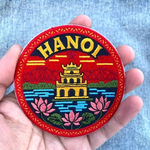 Könnte beinhalten: Ein runder, bestickter Aufnäher mit dem Wort HANOI in gelben Buchstaben am oberen Rand. Das Design zeigt eine gelbe Pagode auf blauem Wasser, umgeben von rosa Lotusblüten und grünen Blättern vor einem leuchtend roten Hintergrund.
