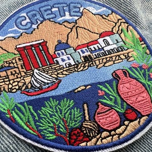 Op de afbeelding: Een ronde geborduurde patch met de tekst CRETE bovenaan. Het ontwerp toont een kustlandschap met ruïnes met rode zuilen, witte gebouwen met blauwe koepels, een zeilboot op blauw water en aardewerk in terracotta-kleur op de voorgrond.