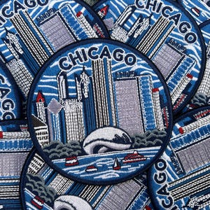 Op de afbeelding: Een ronde geborduurde patch met de skyline van Chicago in blauwe, grijze en witte tinten. Het ontwerp bevat hoge wolkenkrabbers, het Cloud Gate-beeldhouwwerk en boten op het water, met het woord CHICAGO in een boog aan de bovenzijde.