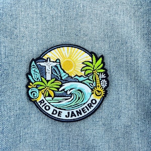 May include: A circular embroidered patch featuring a vibrant Rio de Janeiro scene with the Christ the Redeemer statue, a bright yellow sun, green palm trees, and a blue ocean wave. The text RIO DE JANEIRO is stitched in black along the bottom edge.