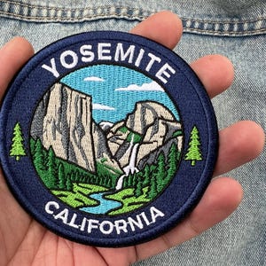 Puede incluir: Un parche bordado circular que muestra un paisaje de Yosemite, California. El diseño incluye acantilados de granito, una cascada, un río azul y pinos verdes sobre un cielo azul claro. El texto YOSEMITE y CALIFORNIA está en color blanco.