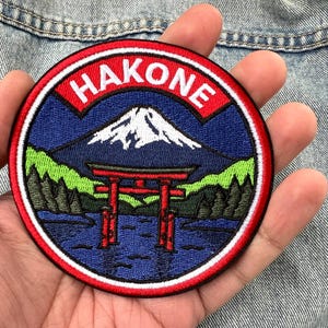 May include: A circular embroidered patch featuring a landscape scene of a snow-capped mountain, a red torii gate in blue water, and green trees. The word HAKONE is stitched in white capital letters on a red arched banner at the top of the patch.