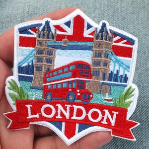 Op de afbeelding: Een schildvormige geborduurde patch met een rode dubbeldekkerbus op de voorgrond, de Tower Bridge op de achtergrond en een Union Jack-vlagmotief. Een rode banner onderaan toont de witte tekst LONDON in hoofdletters.