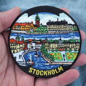 May include: A circular embroidered patch featuring a colourful cityscape of Stockholm, Sweden. The design depicts buildings, a river, and green spaces, with the word STOCKHOLM stitched in yellow capital letters at the bottom on a black border.