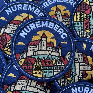 Könnte beinhalten: Eine Sammlung runder gestickter Aufnäher mit blauem Hintergrund und dem weißen Schriftzug NUREMBERG. Das Design zeigt eine zentrale Burg mit roten Dächern und gelben Sonnenakzenten, umgeben von detaillierten architektonischen Gebäudemotiven.