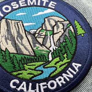 Può includere: Una toppa ricamata circolare che raffigura un paesaggio di Yosemite, in California. Il design include scogliere di granito color marrone chiaro, una cascata bianca, pini verdi e un fiume blu sotto un cielo azzurro. Il testo YOSEMITE CALIFORNIA è bianco.