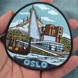 Op de afbeelding: Een ronde geborduurde patch met een gestileerde afbeelding van Oslo, Noorwegen. Het ontwerp bevat een Vikingschip, het stadhuis van Oslo en de Holmenkollen-skischans tegen een lichtblauwe lucht met wolken. Het woord OSLO is in het blauw geborduurd.