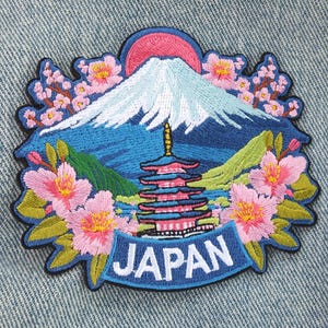 Puede incluir: Un parche bordado que muestra un pico de montaña nevado bajo un sol rosa, una pagoda de varios niveles y flores de cerezo rosas. El diseño incluye una banda azul en la parte inferior con el texto blanco JAPAN en letras mayúsculas.