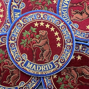 Op de afbeelding: Een ronde geborduurde patch met een bruine beer die op zijn achterpoten staat en naar rode vruchten aan een boom reikt. Het ontwerp bevat zeven gele sterren, een blauwe rand met gouden krulwerk en het woord MADRID in gouden hoofdletters.
