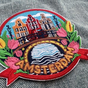 Pode incluir: Um emblema bordado circular apresentando uma cena de Amsterdã com casas de canal coloridas, uma ponte sobre a água e tulipas vibrantes em tons de rosa e amarelo. A palavra AMSTERDAM está bordada em letras douradas numa faixa vermelha.